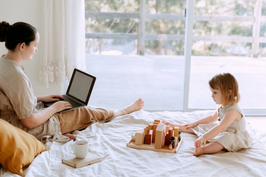 Side view of casual woman sitting on bed and surfing laptop with blank screen while adorable kid playing with toys in cozy bedroom