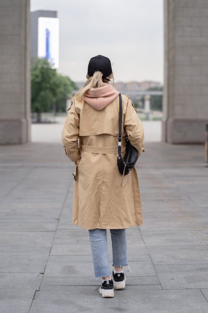 A person wearing a beige coat and jeans walks outdoors, showcasing a casual street fashion style.