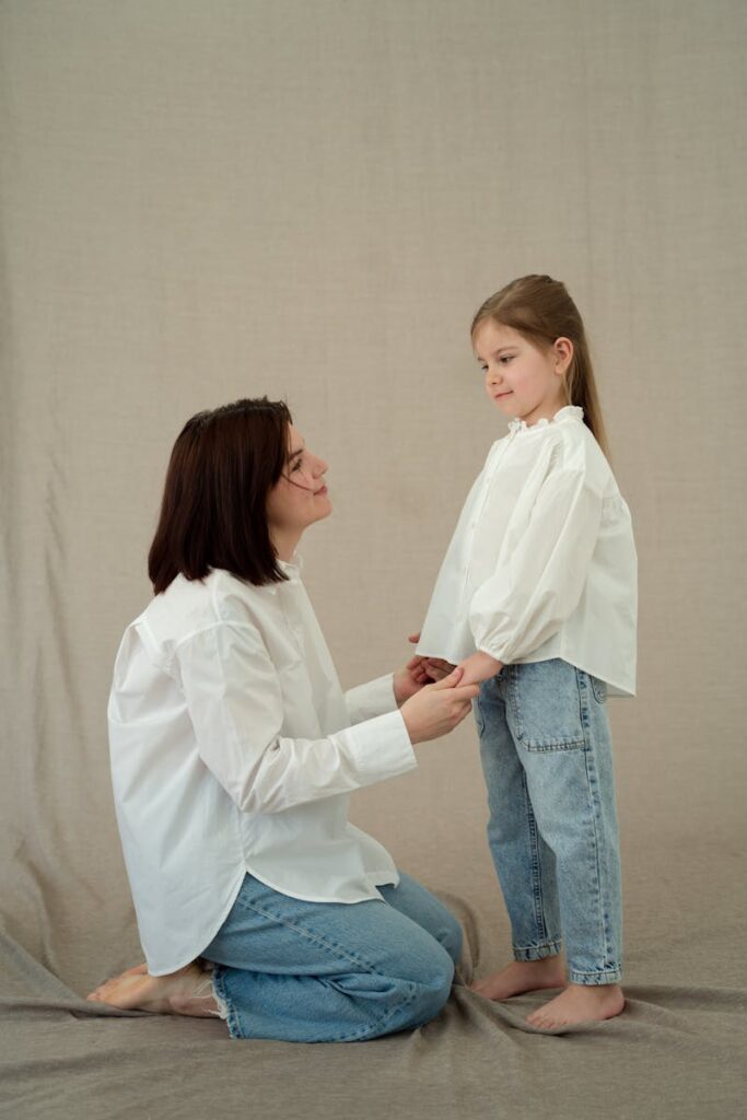 A mother and daughter in matching white tops and jeans share a tender moment indoors, exuding warmth and affection.