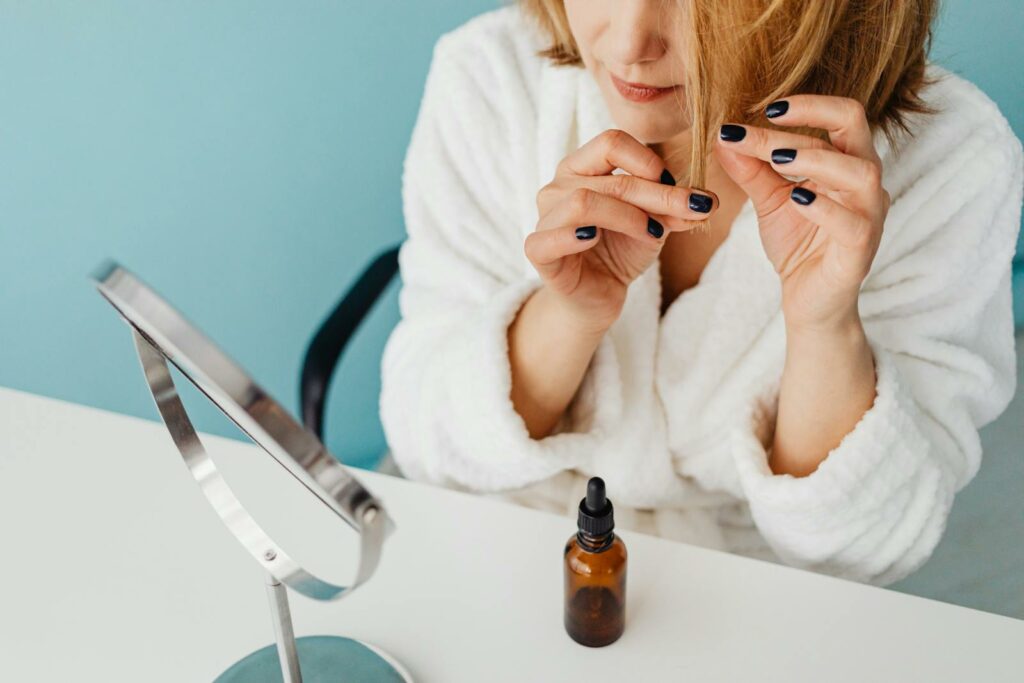 Woman in a bathrobe applying hair oil beside a mirror on a blue background.